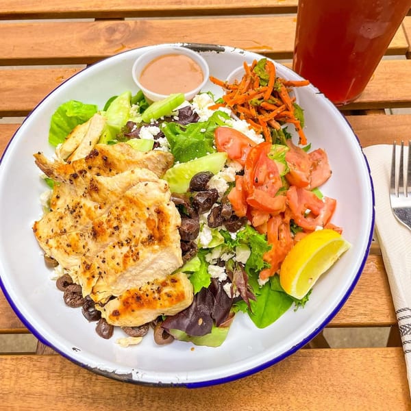 Grilled chicken salad with lettuce, tomatoes, olives, feta, and a lemon wedge on a wooden table.