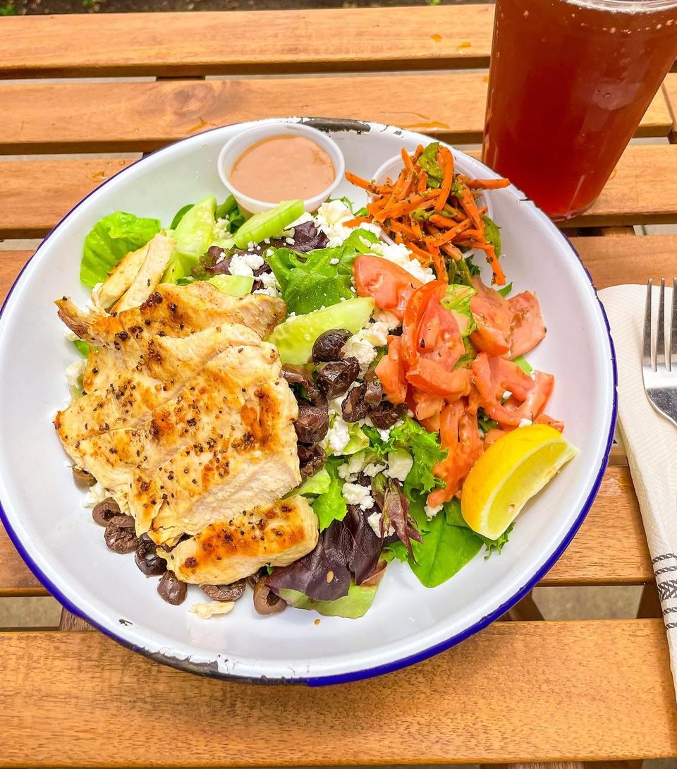 Grilled chicken salad with lettuce, tomatoes, olives, feta, and a lemon wedge on a wooden table.