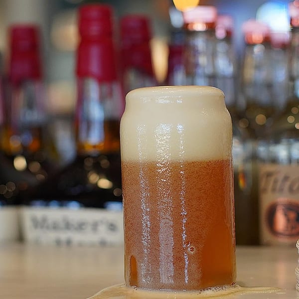 An overflowing glass of beer with a thick head of foam, spilling onto a white surface.
