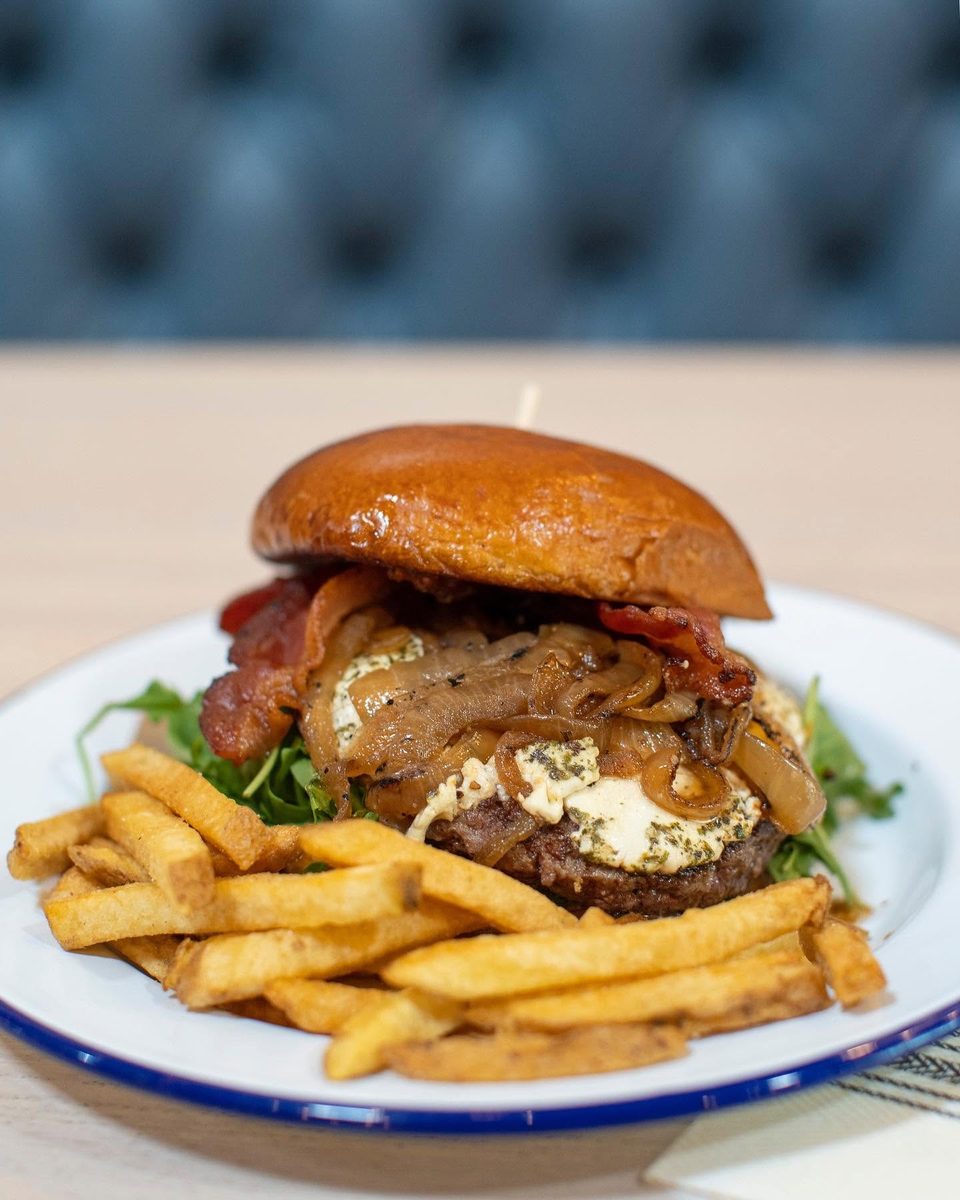 Gourmet burger with bacon, caramelized onions, goat cheese, and fries on a white plate.