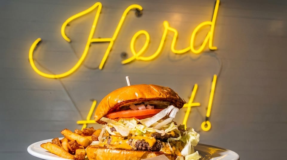 A double cheeseburger with lettuce, tomato, and onion, served with fries, in front of a neon sign.
