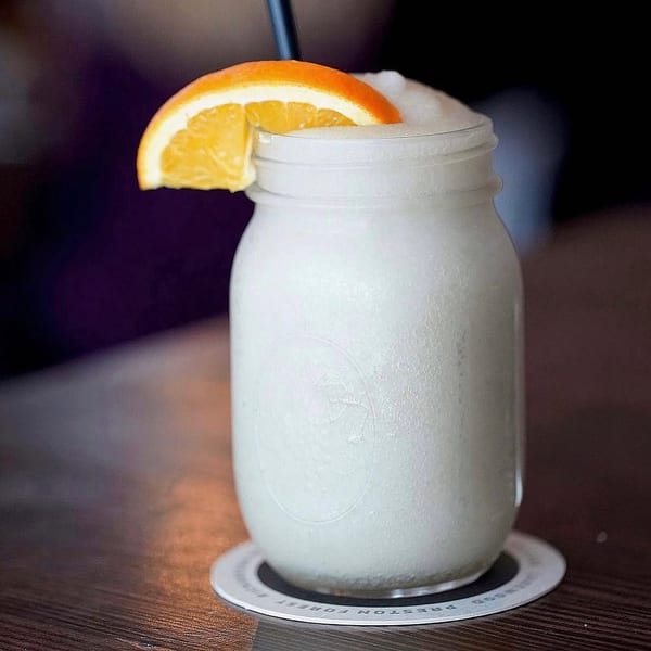 A frosty white drink in a mason jar, garnished with an orange slice and black straw.