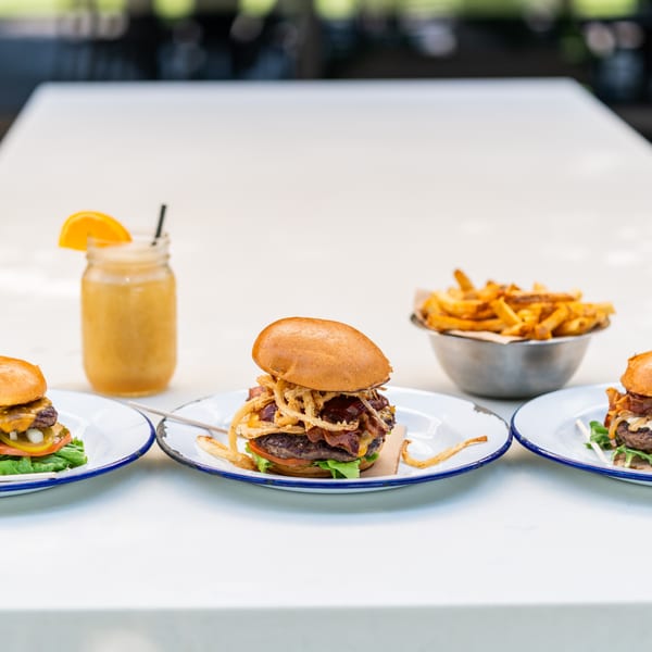 Three gourmet burgers, a glass of orange juice, and a bowl of fries on a white table.