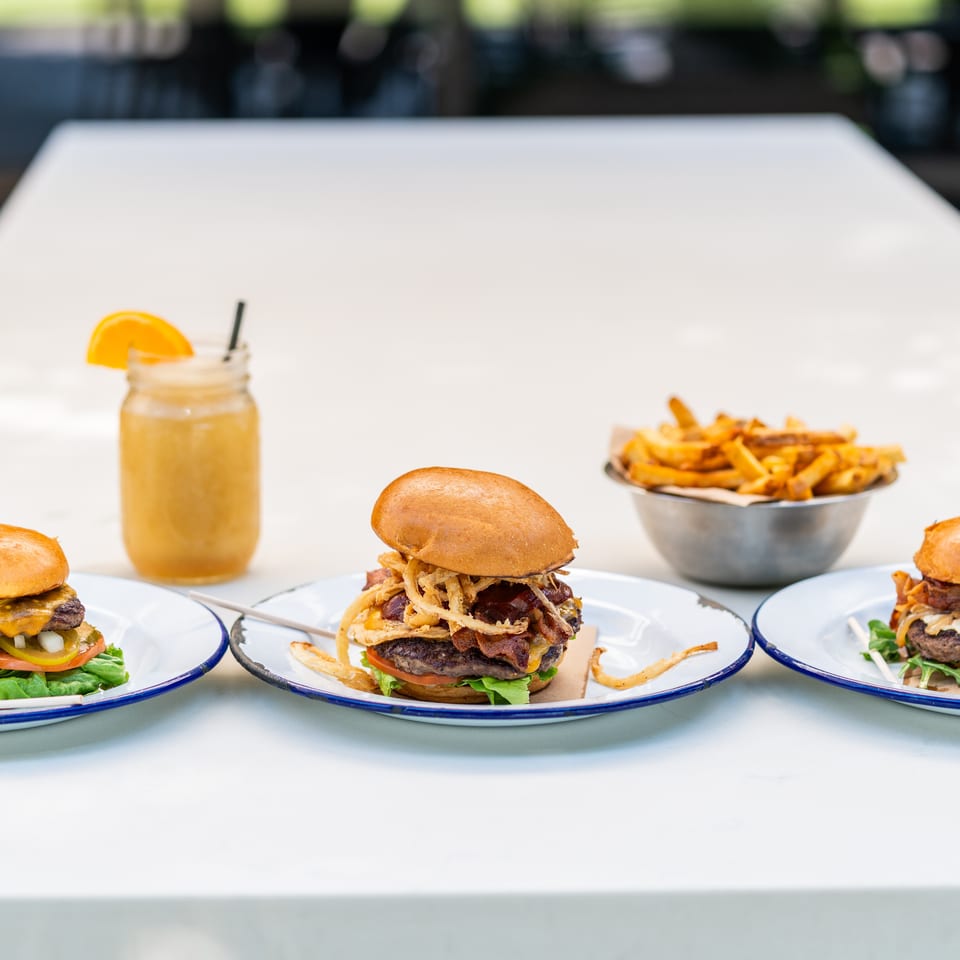 Three gourmet burgers, a glass of orange juice, and a bowl of fries on a white table.