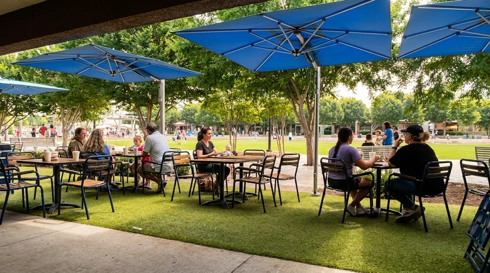 People dine at outdoor tables with blue umbrellas on artificial turf, overlooking a park.