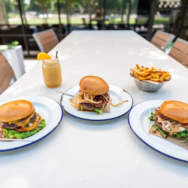 Three gourmet burgers, a side of fries, and a drink on a white outdoor table.