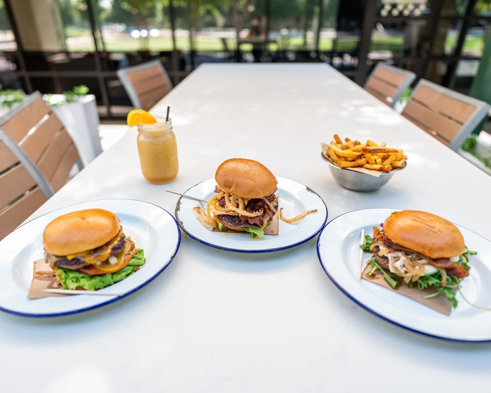 Three gourmet burgers, a side of fries, and a drink on a white outdoor table.