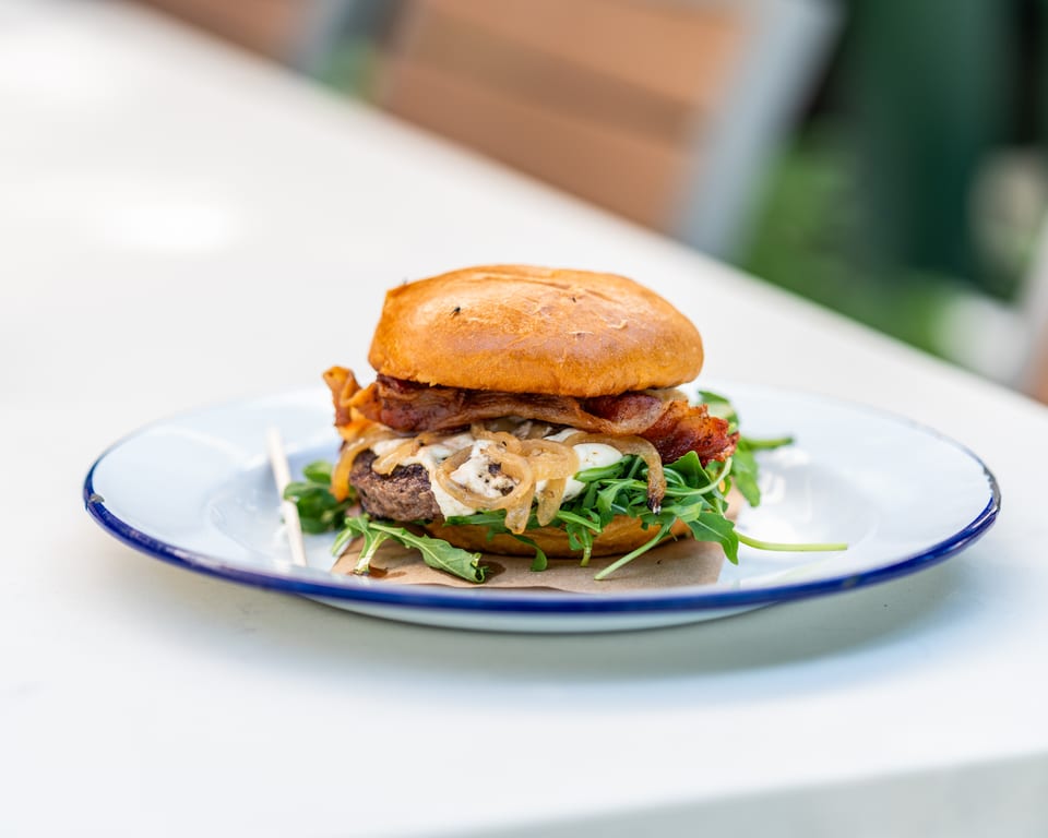 Gourmet burger with bacon, caramelized onions, cheese, and arugula on a white plate.