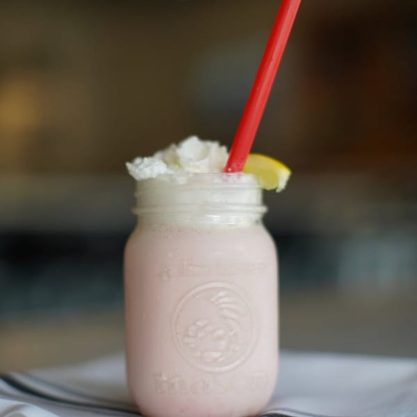 Pink milkshake in a mason jar with whipped cream, a lemon slice, and a red straw.