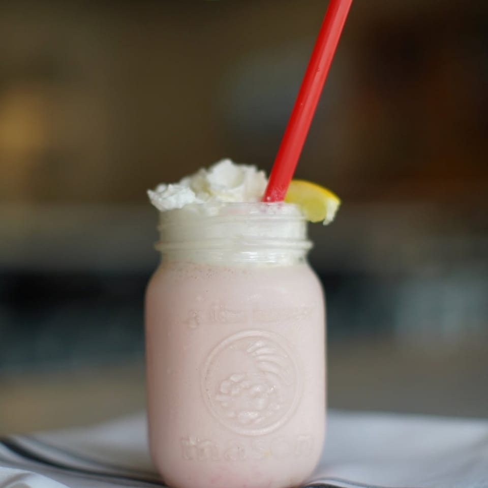 Pink milkshake in a mason jar with whipped cream, a lemon slice, and a red straw.