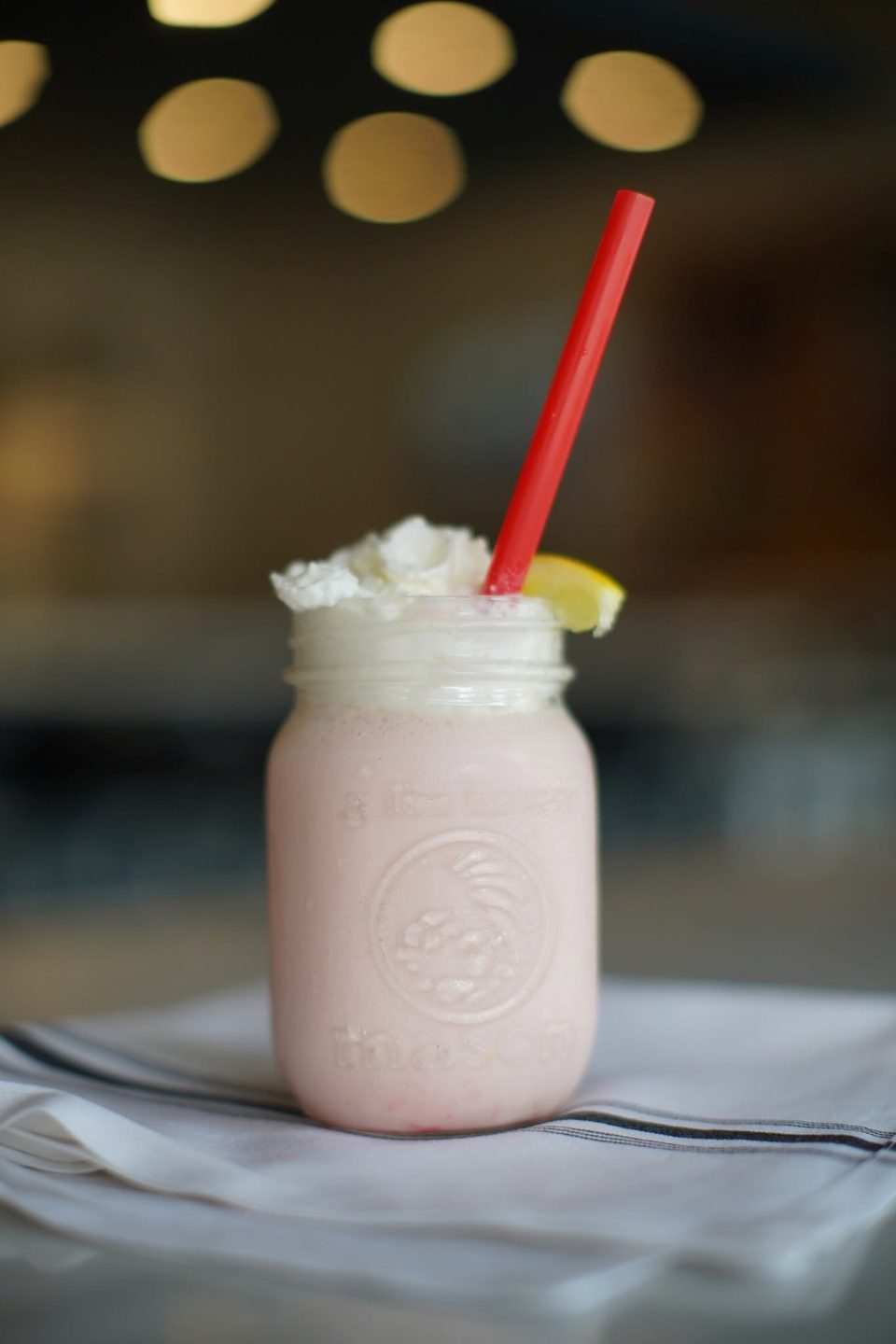 Pink milkshake in a mason jar with whipped cream, a lemon slice, and a red straw.