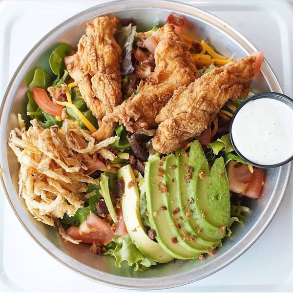 Crispy chicken tender salad with avocado, fried onions, tomatoes, cheese, bacon, and ranch dressing.