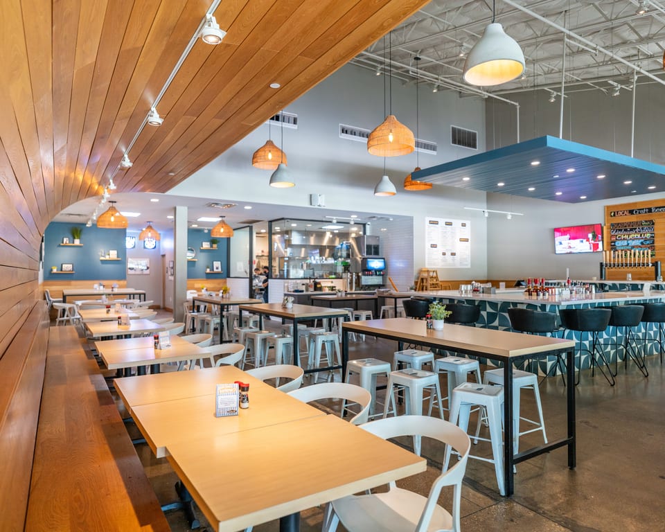 A modern restaurant interior with light wood tables, benches, and a bar with blue patterned tiles.
